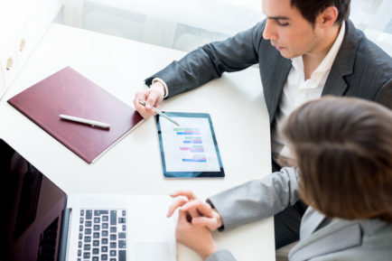 Top view of two business people, a man and woman, analysing a business graph on a tablet computer as they sit at the office desk.