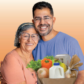 Image of hispanic man standing next to his elderly mother smiling and holding a brown paper bag with fresh grocery items.