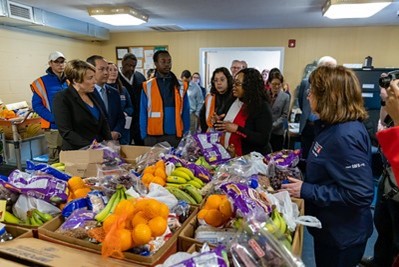 Governor Maura Healey at the Dwelling House of Hope in Lowell Massachusetts, with Executive Director Levenia Furusa speaking to her and a group in front of a table filled with boxes of fresh produce.